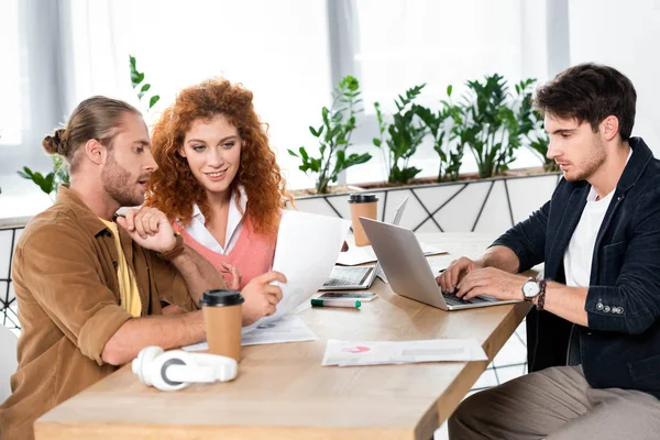 Tres amigos haciendo papeleo y usando portátil en la oficina - foto de stock