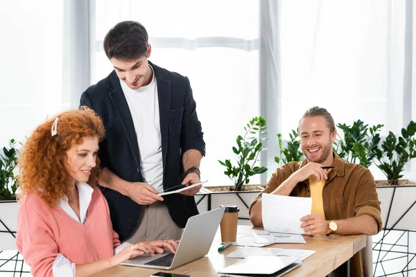 Tres amigos usando el ordenador portátil, sosteniendo la tableta digital y haciendo papeleo - foto de stock