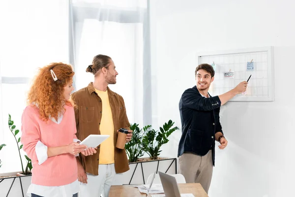 Schöner und lächelnder Mann zeigt auf Whiteboard und schaut seine Freunde an — Stockfoto