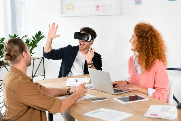 Hombre de negocios sonriente con realidad virtual mostrando manos y amigos mirándolo - foto de stock