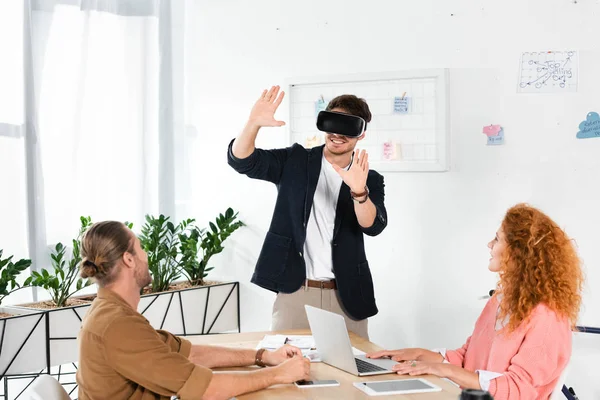 Homem de negócios sorridente com realidade virtual mostrando as mãos e amigos olhando para ele — Fotografia de Stock