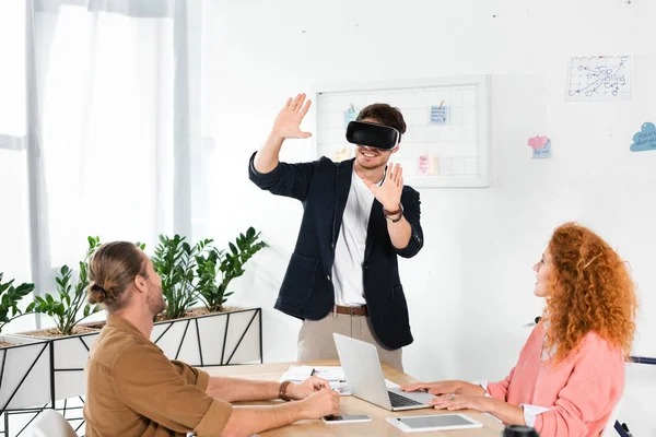 Hombre de negocios sonriente con realidad virtual mostrando manos y amigos mirándolo - foto de stock