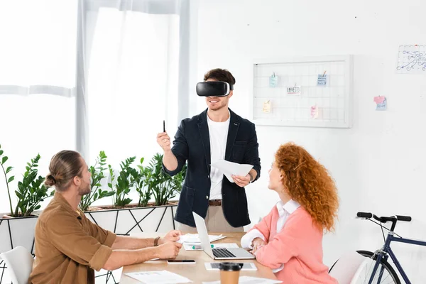Smiling businessman with virtual reality headset holding pen and papers and friends looking at him — Stock Photo