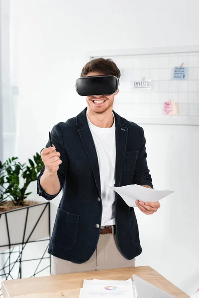 Hombre de negocios sonriente en camisa con auriculares de realidad virtual con bolígrafo y papeles - foto de stock