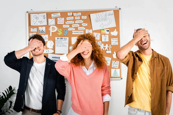 Tres amigos sonrientes oscureciendo caras con las manos en la oficina - foto de stock