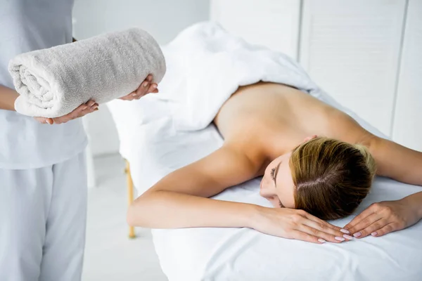 Vista recortada de masajista sosteniendo toalla y mujer acostada en alfombra de masaje - foto de stock