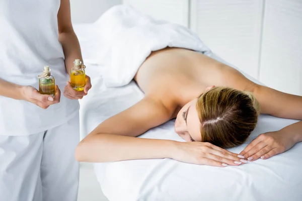 Vista cortada de massagista segurando óleos de fragrância e mulher deitada no tapete de massagem — Fotografia de Stock