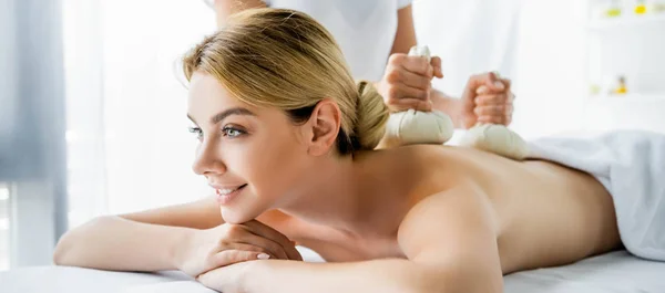 Plano panorámico de masajista haciendo masaje de espalda con bolas de hierbas a mujer sonriente en spa - foto de stock