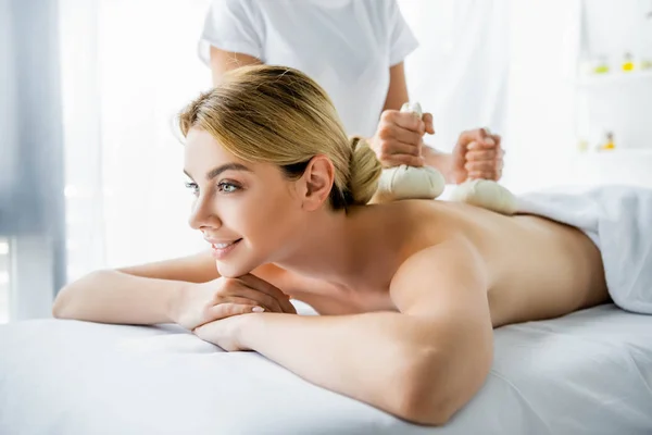 Vista cortada de massagista fazendo massagem nas costas com bolas de ervas para mulher sorridente no spa — Fotografia de Stock