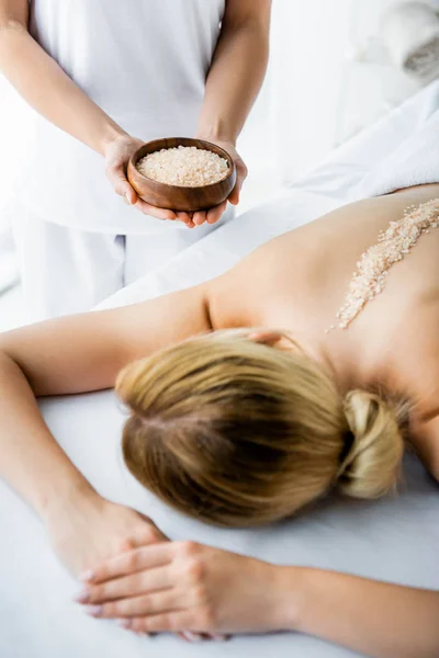 Vista ritagliata della ciotola massaggiatore con sale marino e donna sdraiata sul tappeto da massaggio in spa — Foto stock