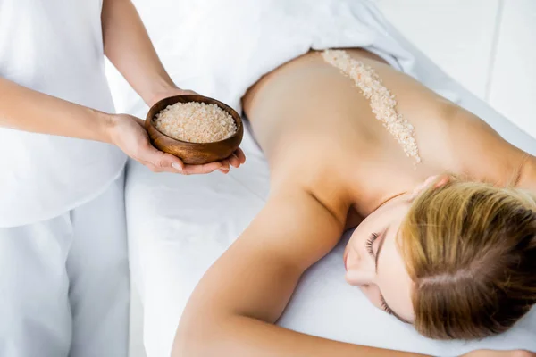 Обрізаний вид на масажиста, що тримає миску з морською сіллю і жінку, що лежить на масажному килимку в спа-центрі — стокове фото