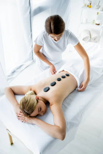Vista de ángulo alto de masajista haciendo masaje de piedra caliente a la mujer en el spa - foto de stock