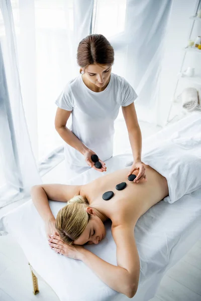 Высокий угол зрения массажиста, делающего массаж горячими камнями женщине в спа — стоковое фото