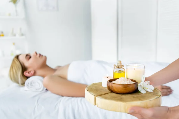 Selective focus of masseur holding board with fragrance oil, candles, sea salt and flower — Stock Photo