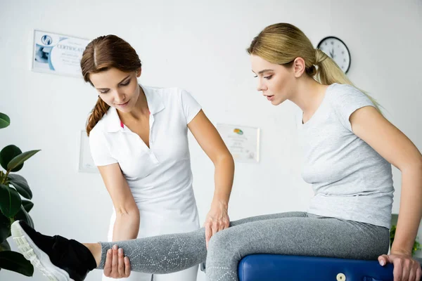 Chiropraktiker berührt Bein einer attraktiven Patientin im grauen T-Shirt — Stockfoto