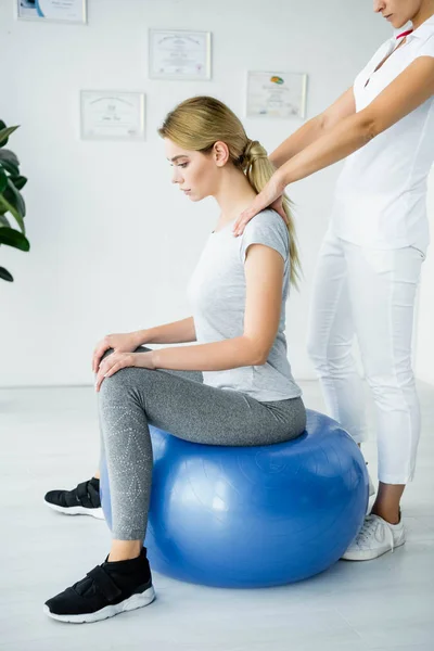 Chiropraktiker berührt Schultern attraktiver Patientin auf blauem Trainingsball — Stockfoto