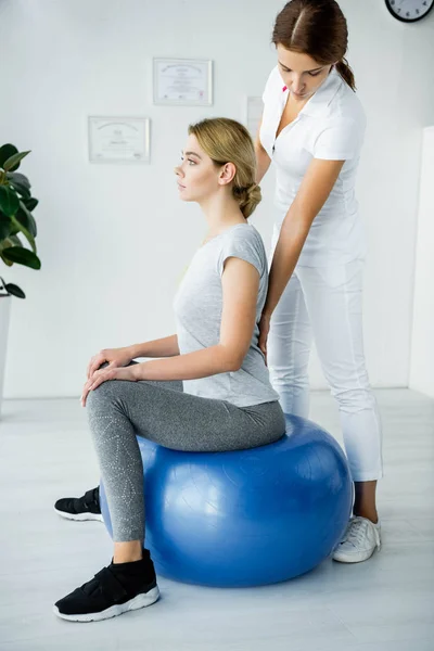 Atractiva paciente sentada en bola de ejercicio azul y quiropráctico tocando su espalda - foto de stock