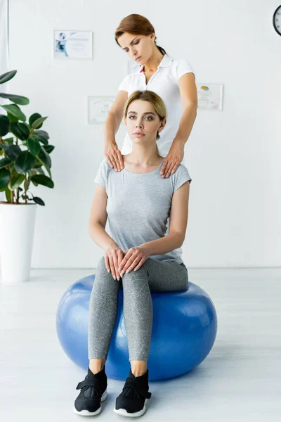 Привлекательная пациентка сидит на синем мяче для упражнений и хиропрактик трогает ее за плечи — стоковое фото