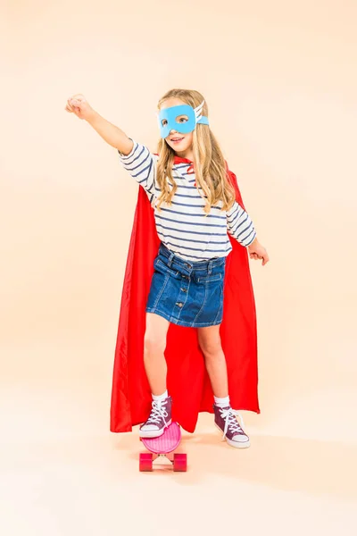 Vue pleine longueur de l'enfant dans le masque et le manteau de héros tenant le poing vers le haut tout en se tenant sur skateboard sur rose — Photo de stock