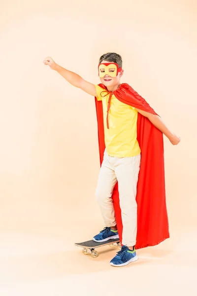 Full length view of child in mask and hero cloak holding fist up while standing in skateboard on pink — Stock Photo