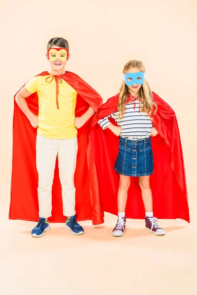 Full length view of two smiling kids in masks and red cloaks standing with hands on hips on pink — Stock Photo