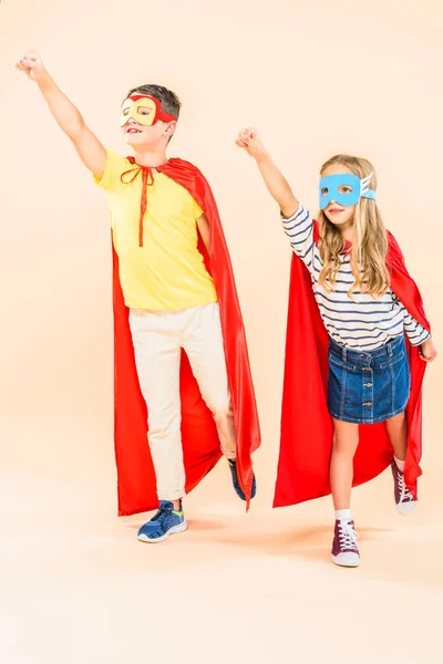 Vista completa de dos niños sonrientes con máscaras y capas rojas sosteniendo puños en rosa - foto de stock