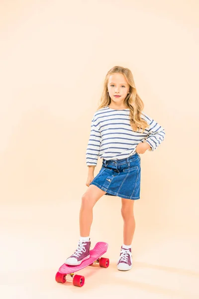 Vue pleine longueur de l'enfant debout sur skateboard avec la main sur la hanche sur rose — Photo de stock