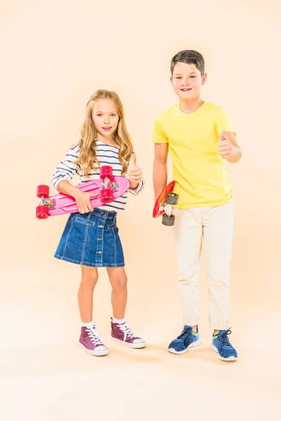 Vue pleine longueur de deux enfants souriants tenant des planches à roulettes et montrant pouces vers le haut sur rose — Photo de stock