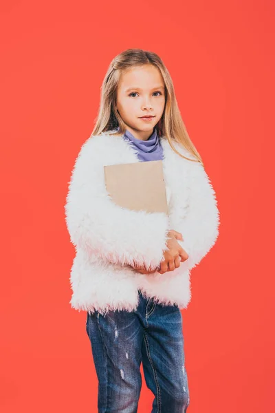 Kid in autumn outfit holding book and looking at camera isolated on red — Stock Photo