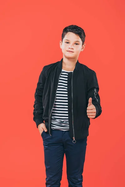 Vista frontal da criança de jaqueta de pé com a mão no bolso e mostrando o polegar para cima isolado no vermelho — Fotografia de Stock