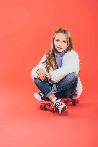 Smiling kid in autumn outfit sitting on skateboard on red — Stock Photo