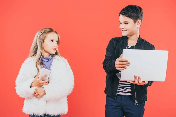 Smiling kid with smartphone and laptop looking at each other isolated on red — Stock Photo