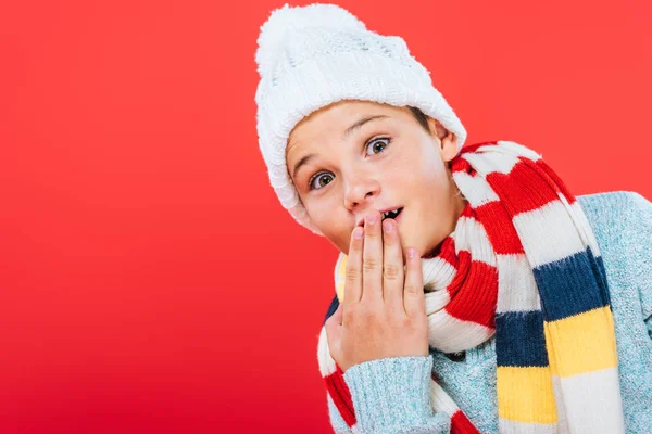 Niño sorprendido en sombrero y bufanda que cubre la boca con la mano aislada en rojo - foto de stock