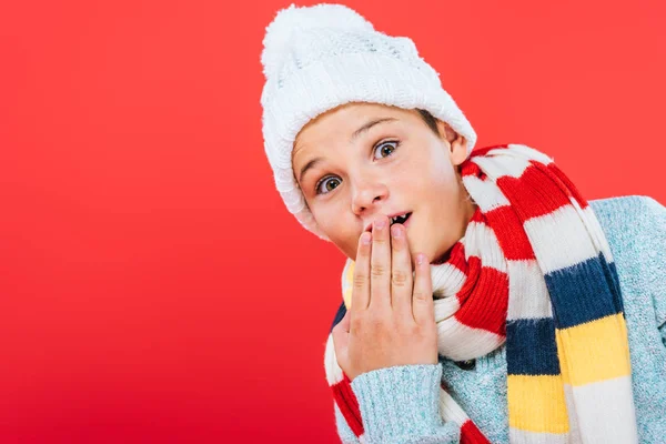 Bambino sorpreso in cappello e sciarpa che copre la bocca con mano isolata su rosso — Foto stock