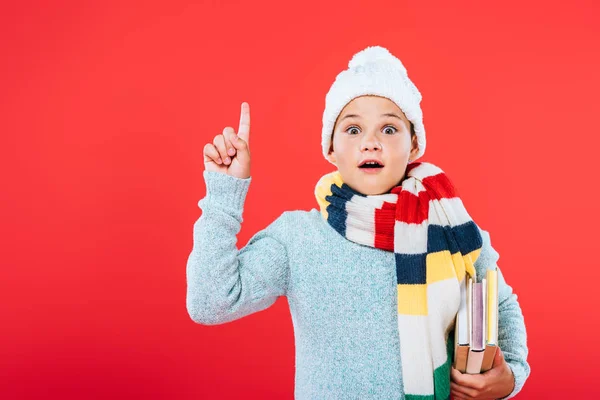 Front view of shocked kid in hat and scarf holding books and showing idea sign isolated on red — Stock Photo