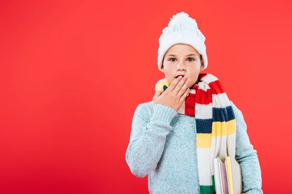 Front view of surprised kid in hat and scarf covering mouth with hand isolated on red — Stock Photo