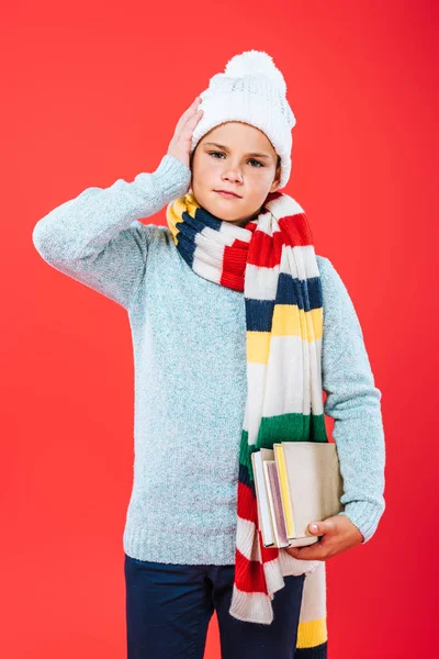 Vista frontal de niño pensativo en sombrero y bufanda sosteniendo libros aislados en rojo - foto de stock