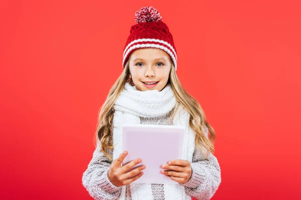 Vue de face de l'enfant souriant en chapeau et écharpe à l'aide d'une tablette numérique isolée sur rouge — Photo de stock