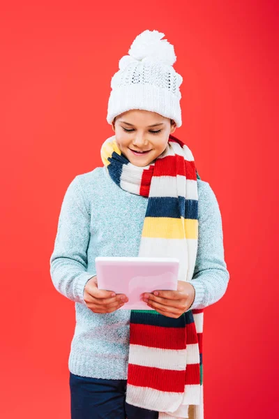 Front view of smiling child in hat and scarf using digital tablet isolated on red — Stock Photo