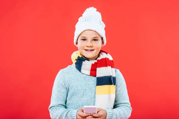 Front view of smiling kid in hat and scarf using smartphone isolated on red — Stock Photo