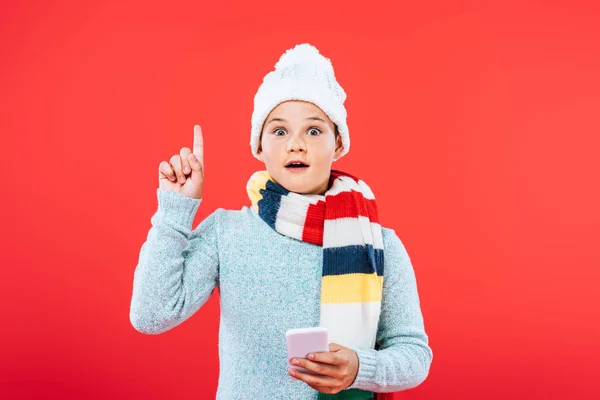 Вид спереду шокованої дитини в капелюсі і шарфі, використовуючи смартфон і показуючи знак ідеї ізольовано на червоному — стокове фото