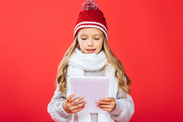 Vista frontale del bambino sorridente in cappello e sciarpa con tablet digitale isolato sul rosso — Foto stock