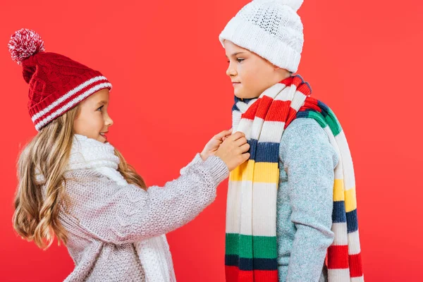 Two kids in hats and scarfs looking at each other isolated on red — Stock Photo