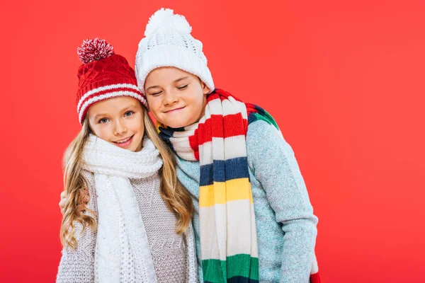 Два улыбающихся ребенка в шляпах и шарфах, обнимающие изолированных на красный — стоковое фото