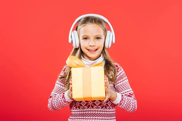 Vorderansicht eines lächelnden Kindes im Pullover, das mit Kopfhörern Musik hört und Geschenke isoliert auf Rot hält — Stockfoto