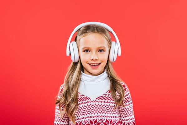 Front view of smiling kid in sweater listening music in headphones isolated on red — Stock Photo
