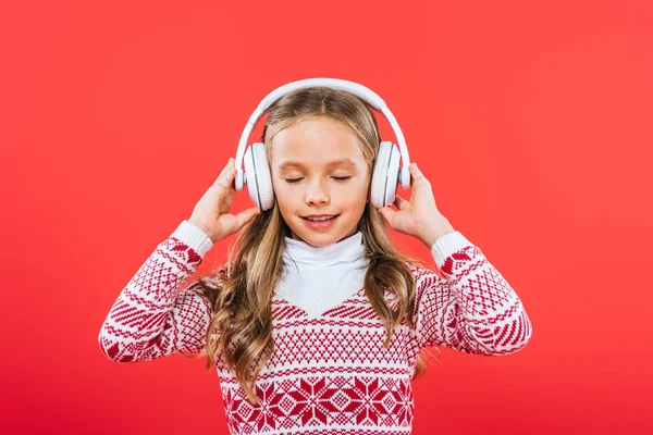 Vista frontale del bambino sorridente in maglione ascoltare musica in cuffie isolate sul rosso — Foto stock