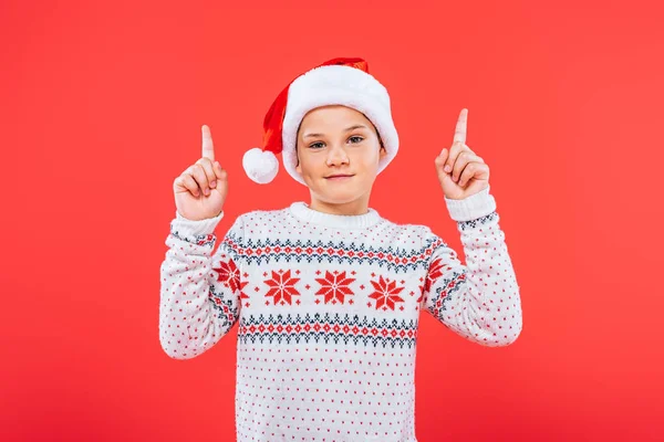 Front view of smiling kid in sweater and santa hat isolated on red — Stock Photo