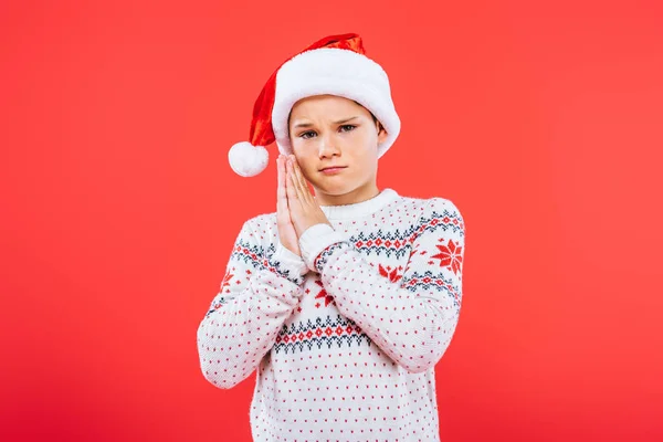 Triste enfant en pull et chapeau santa montrant s'il vous plaît geste isolé sur rouge — Photo de stock