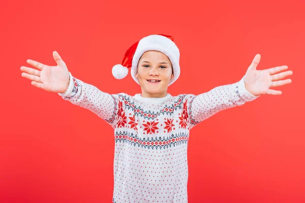 Vorderseite des lächelnden Kindes in Pullover und Weihnachtsmütze isoliert auf rot — Stockfoto